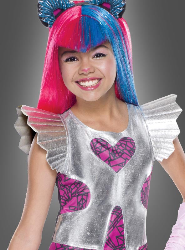 Ein Mädchen im „Catty Noir“-Kostüm von Monster High – bestehend aus einem silbernen Oberteil mit Herzmustern, Rüschen an den Schultern und leuchtend pink-blauen Haaren – lächelt vor einem grauen Hintergrund.