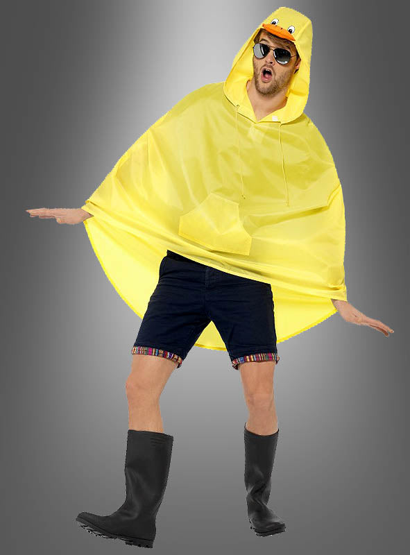 Eine Person, die einen gelben „Regencape Ente“-Umhang mit einer Kapuze in Entenform, schwarze Shorts, schwarze Stiefel und eine Sonnenbrille trägt, steht auf einem Bein vor einem grauen Hintergrund.