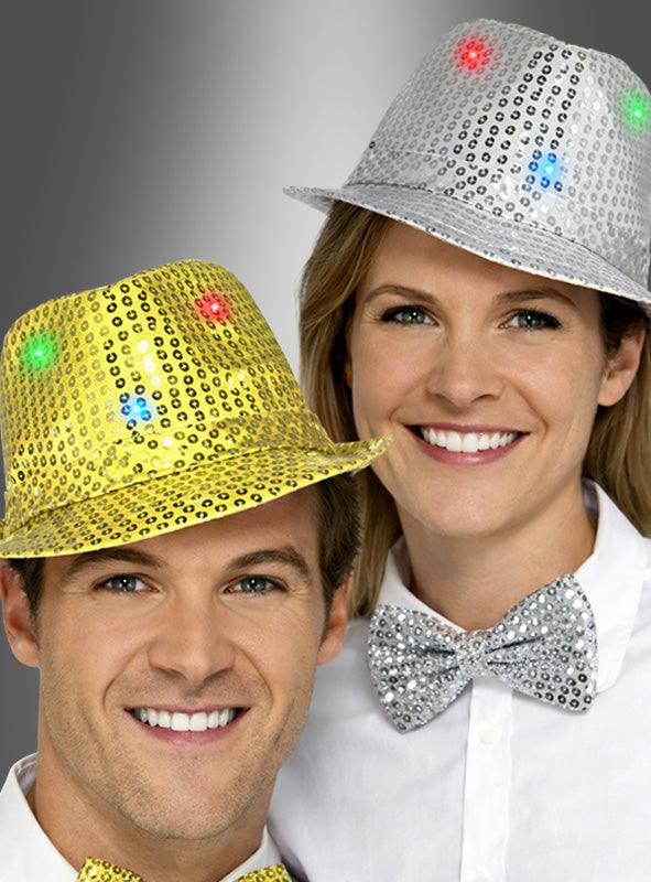 Zwei Personen, die glitzernde Trilby-Hüte mit LED in Silber oder Gold tragen – ideal für Partys oder als klassisches Karnevalsaccessoire – sowie dazu passende Pailletten-Fliegen, lächeln strahlend vor einem grauen Hintergrund mit Farbverlauf.