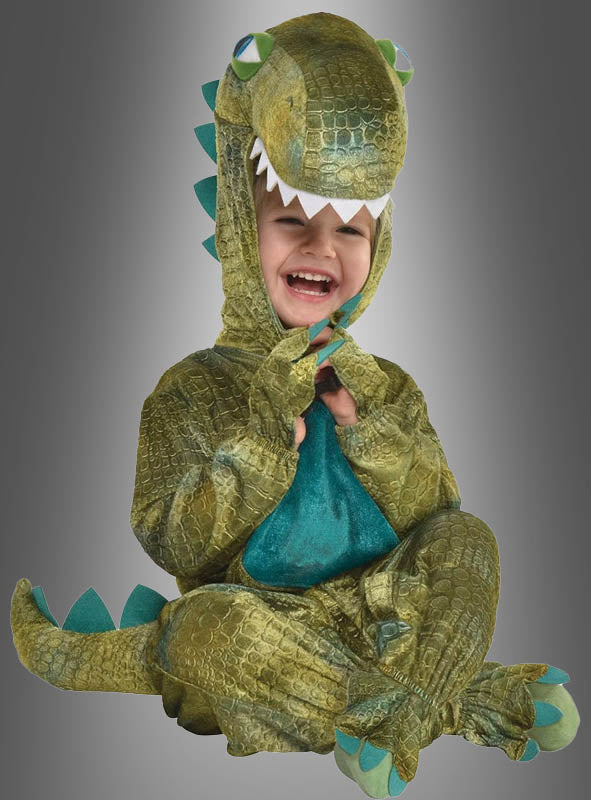 Ein Kind sitzt lächelnd vor einem grauen Hintergrund und trägt das „Dino-Kostüm für Kleinkinder“, ein grünes Kostüm mit einer Kapuze in Form eines Dinosaurierkopfes – perfekt für den Kinderfasching.