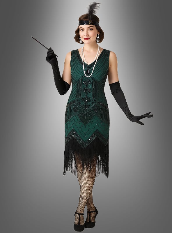 Eine Frau präsentiert das „20er-Jahre-Kleid Shirley“ mit Accessoires in Grün – bestehend aus einem grünen Charleston-Kleid, schwarzen Handschuhen, Netzstrümpfen, Perlen, einem Haarreif mit Federn und einer Zigarettenspitze –, und fängt so vor einem grauen Hintergrund die Essenz der klassischen Accessoires der „Roaring Twenties“ ein.