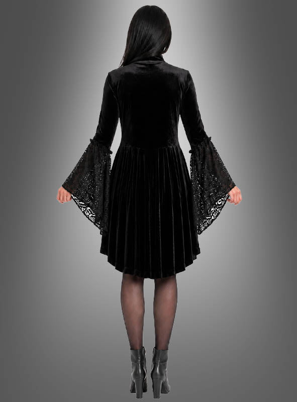 Eine Frau, die das Gothic-Kleid „Lucy mit Spitze“ aus schwarzem Samt mit ausgestellten Trompetenärmeln trägt und dazu Stiefeletten kombiniert hat, steht vor einem grauen Hintergrund mit Farbverlauf und wendet der Kamera den Rücken zu.