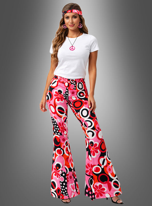 Mit der Schlaghose mit Accessoires „Dream Circles“ in Rosa-Rot – einem weißen T-Shirt, einer bunten Hose mit Retro-Muster, einem passenden Stirnband, einer Halskette mit Peace-Zeichen und Ohrringen – eignet sich dieser Look perfekt für jedes Hippie-Kostüm oder jede Woodstock-Party. Vor einem schlichten grauen Hintergrund.