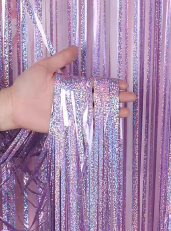 Eine Hand hält Stränge aus glänzendem, schillerndem, lavendelfarbenem Lametta Vorhang 200–250 cm Party-Deko vor einem passenden Hintergrund.