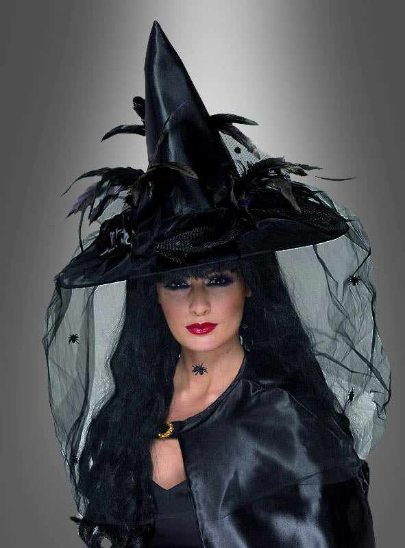 Eine Person in einem schwarzen Hexenkostüm mit dem „Deluxe Hexenhut schwarz mit Federn“ – einem spitzen, mit Federn verzierten Hut –, einem schwarzen Schleier und dunklem Make-up posiert vor einem grauen Hintergrund mit Farbverlauf.
