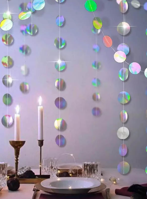 Vor einer Wand, die mit hängenden schillernden Scheiben und den effektvollen Hologramm-Girlanden „400 cm Sterne oder Kreise“ geschmückt ist, steht ein Esstisch mit weißen Tellern und Kerzen, der der Partydekoration eine festliche Note verleiht.