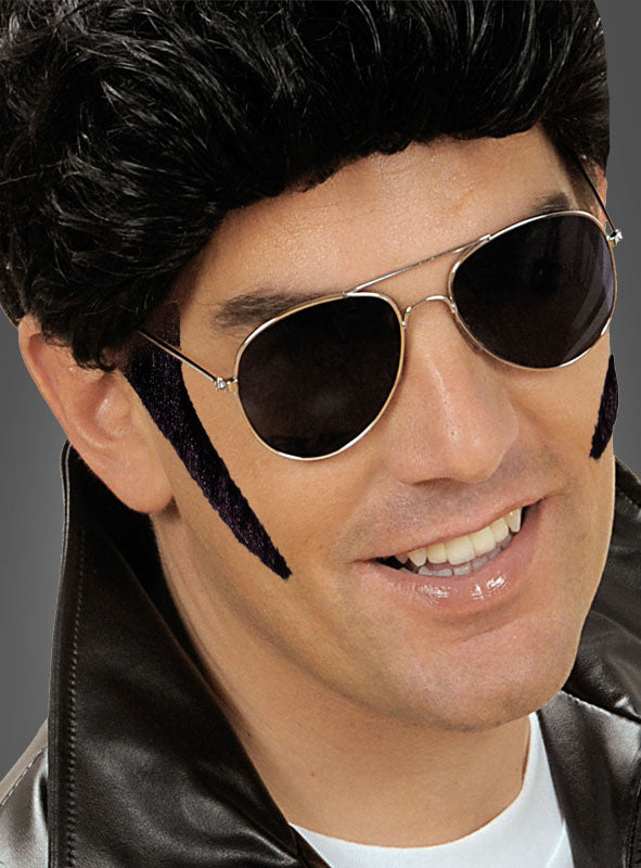 Ein Mann trägt eine schwarze Perücke, eine Pilotenbrille, eine schwarze Lederjacke und „Koteletten zum Ankleben“ als Kostümaccessoires und lächelt in die Kamera.