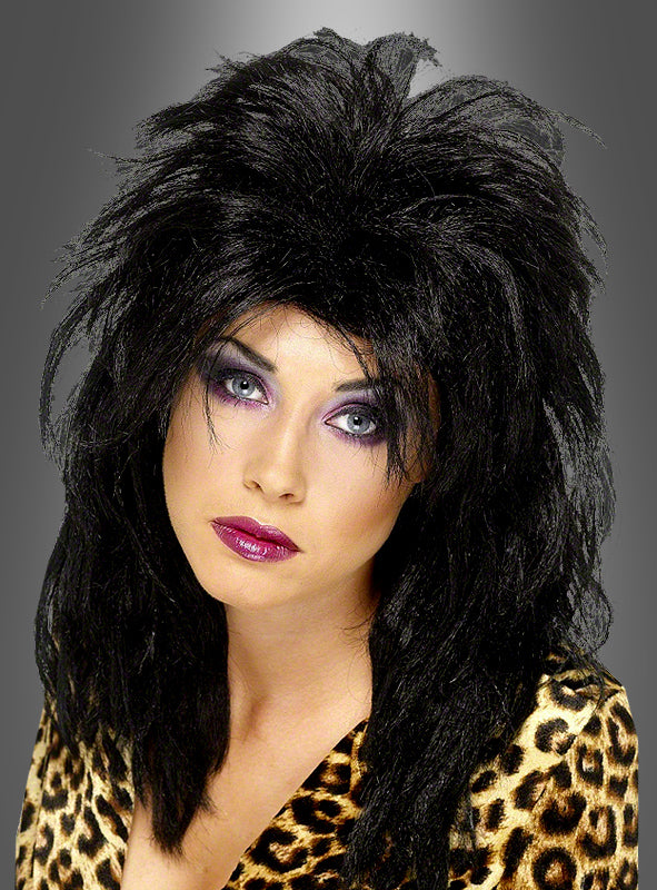 Eine Person mit toupiertem schwarzem Haar, das zu einer 80er-Jahre-Pop-Sternchen-Perücke gestylt ist, und auffälligem lila Make-up, die ein Oberteil mit Leopardenmuster trägt, posiert vor einem grauen Hintergrund mit Farbverlauf.
