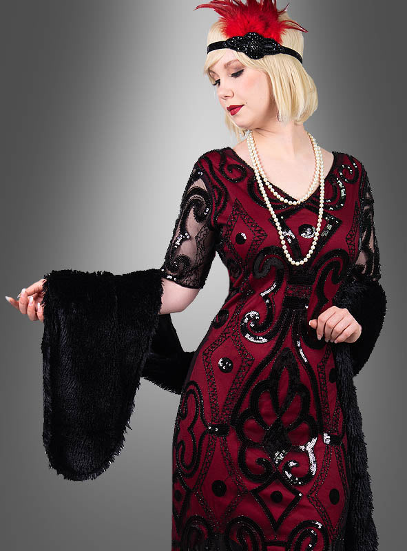 Eine Frau in einem rot-schwarzen Flapper-Kleid mit Pailletten, umhüllt von der „Lange Stola 20er Jahre“ in klassischem Schwarz als elegante Vintage-Stola, ergänzt durch eine lange Perlenkette und ein rotes Stirnband mit Federn – ihr Look wird durch Charleston-Kostüm-Accessoires perfekt abgerundet –, vor einem schlichten grauen Hintergrund.