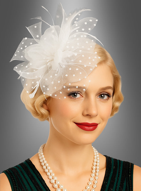 Eine Frau mit kurzen blonden Haaren trägt einen weißen, mit Polka-Dots verzierten Fascinator-Minihut mit Tüll als elegantes Accessoire, knallroten Lippenstift und eine zweireihige Perlenkette und posiert vor einem grauen Hintergrund.