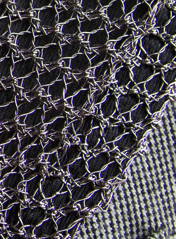 Nahaufnahme eines Metalldrahtgeflechts, das einen strukturierten schwarzen und grauen Stoffhintergrund überlagert und an die filigranen Details eines Ritterkostüms für Kinder erinnert.