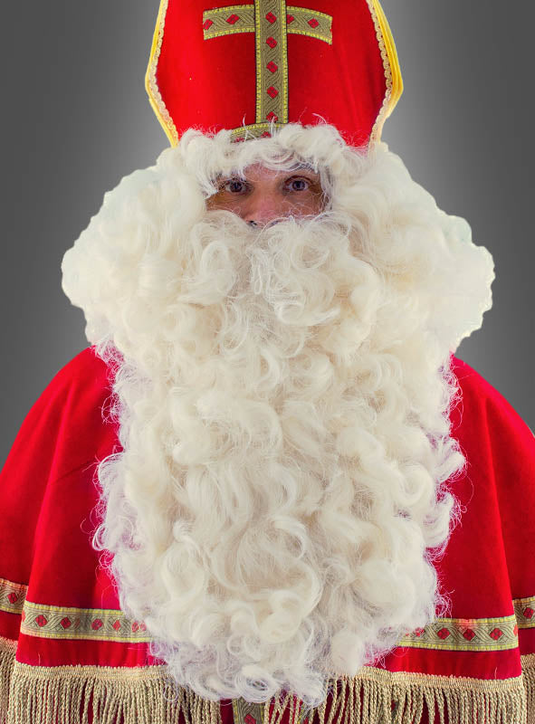 Eine Person, die als Nikolaus in einem rot-goldenen Gewand und einer Mütze verkleidet ist und den „Deluxe Bischof Nikolaus Bart und Perücke“ trägt, steht vor einem schlichten grauen Hintergrund.