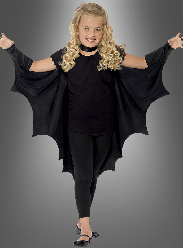 Vor einem grauen Hintergrund steht ein kleines Mädchen, das den „Fledermaus Vampir Umhang für Kinder“ trägt, die Flügel ausgebreitet, dazu schwarze Leggings und Schuhe – eine bezaubernde Wahl für Kinderkostüme oder Halloween-Spaß.
