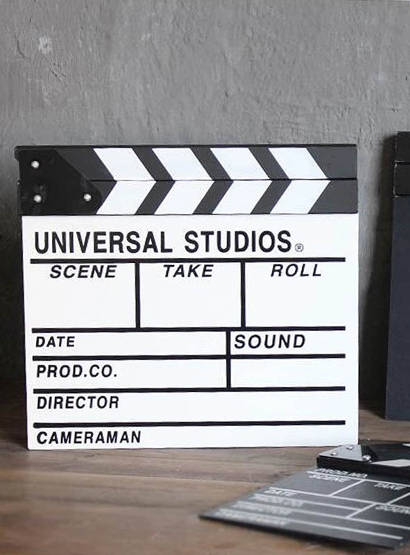 Auf einer Holzunterlage liegt eine schwarz-weiße Filmklappe als Hollywood-Partyzubehör mit Feldern für Szene, Take, Rolle, Datum, Ton, Produktion, Regisseur und Kameramann – damit ist sie das perfekte Kostümzubehör für jede Hollywood-Party.