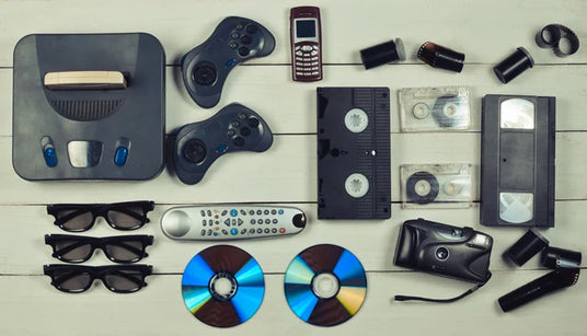 Verschiedene Vintage-Elektronikgeräte, darunter eine Spielkonsole, Controller, ein altes Telefon, VHS-Kassetten, CDs, 3D-Brillen, eine Fernbedienung, Filmdosen und eine Kompaktkamera, angeordnet auf einer Holzunterlage.