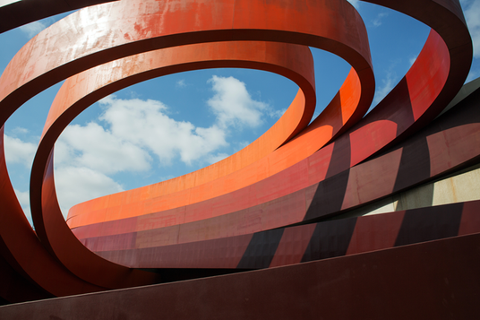 Große rote Metallskulptur in Spiralform, durch deren Windungen der offene Himmel und Wolken zu sehen sind. Das abstrakte Design wirft Schatten auf die Struktur.