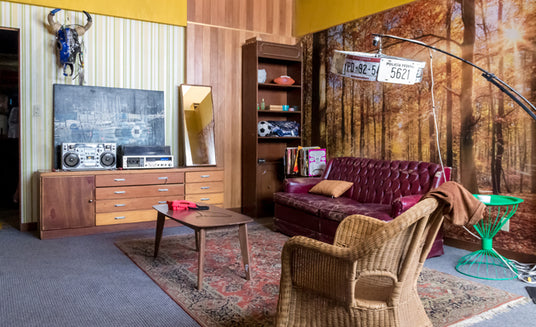 Wohnzimmer mit einem kastanienbraunen Sofa, einem Korbstuhl, einem Holztisch, einer Kommode mit Stereoanlage, einem hohen Regal und einem Wandbild, das einen Herbstwald zeigt. Ein Teppich auf dem Teppichboden, verschiedene Dekoartikel und eine große Lampe in der Ecke.