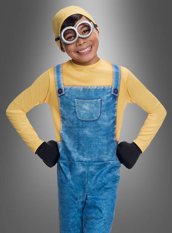 Ein Kind lächelt, während es das Minion-Kostüm „Bob“ für Kinder trägt, bestehend aus einer gelben Mütze, einer runden Schutzbrille, einem gelben Hemd, einer Jeanslatzhose und schwarzen Handschuhen – damit wird der Charme der beliebten Filmfiguren perfekt eingefangen.