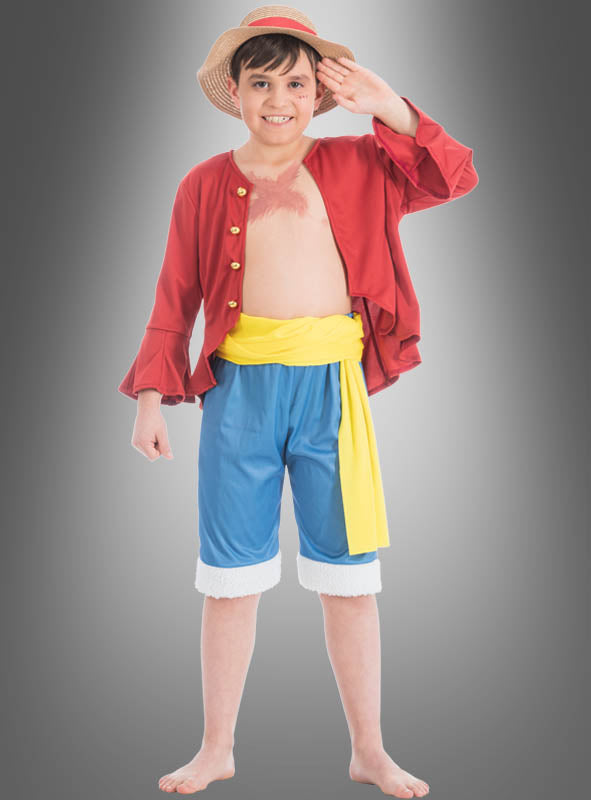 Ein kleiner Junge mit Strohhut, roter Jacke, blauen Shorts und gelber Schärpe steht barfuß da und lächelt, während er vor einem grauen Hintergrund mit Farbverlauf salutiert – ideal für alle, die auf der Suche nach dem perfekten Ruffy-Kostüm für Kinder aus „One Piece“ sind.