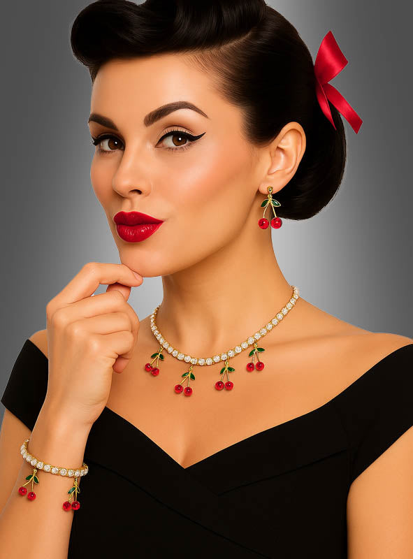 Eine Frau mit einer Retro-Frisur und einer roten Schleife trägt das Strass-Armband „mit Kirschen“ in Gold sowie dazu passende Ohrringe und eine Halskette mit Kirschmotiv. Sie hat einen geschwungenen Eyeliner, roten Lippenstift und ein schwarzes, schulterfreies Oberteil vor einem grauen Hintergrund.