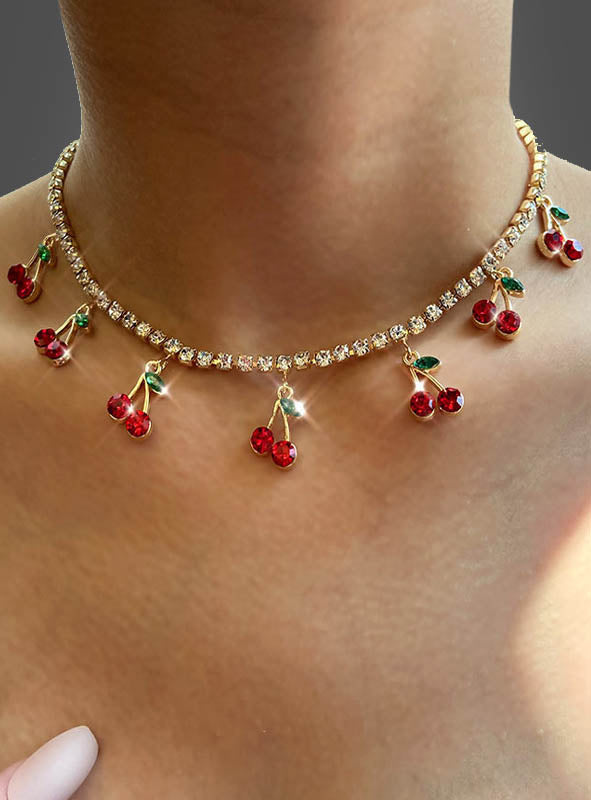 Eine Nahaufnahme einer Person, die die „Strass Kette mit Kirschen 50er“ trägt, eine goldene Halskette, die mit roten Kirschanhängern und klaren, runden Edelsteinen verziert ist.