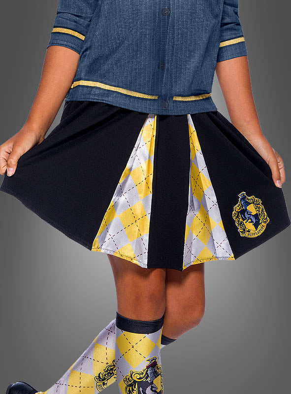 Ein Kind trägt ein blaues Hemd und den „Hufflepuff Rock für Kinder Harry Potter“, einen schwarz-gelben Faltenrock mit Argyle-Muster und offiziellem Wappen, dazu passende Socken – perfekt für alle Fans, die eine authentische Hufflepuff-Schuluniform suchen, die vom Harry-Potter-Kostümstil inspiriert ist.