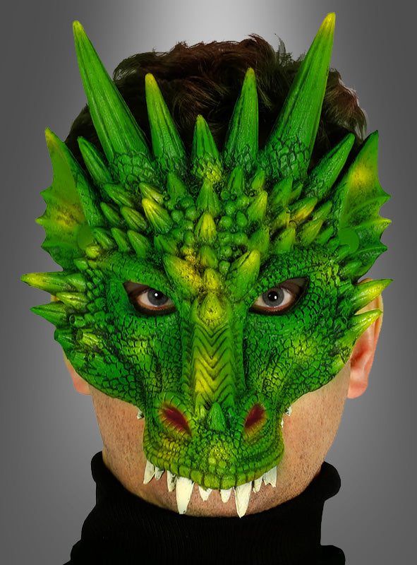 Eine Person, die die „Drachenmaske für Erwachsene“ trägt – eine detailreiche grüne Drachenmaske mit Schuppen, Stacheln und Hörnern, die das gesamte Gesicht bis auf die Augen und den Mund bedeckt –, vor einem schlichten grauen Hintergrund mit Farbverlauf.