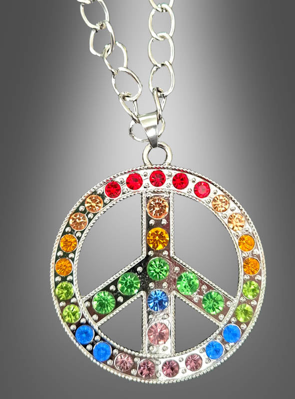 Die „Peace-Kette mit Strass 6 cm“ besteht aus einem silbernen Peace-Zeichen-Anhänger, der mit bunten Strasssteinen verziert ist, an einer Kette hängt und vor einem grauen Hintergrund präsentiert wird.