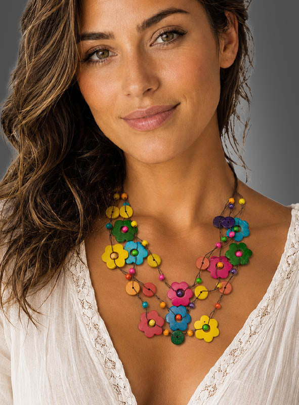 Eine Frau mit welligem braunem Haar, die ein weißes Oberteil trägt und die „Bunte Blumenkette aus Naturmaterial“ um den Hals hat, blickt vor einem neutralen Hintergrund mit einem leichten Lächeln in die Kamera.
