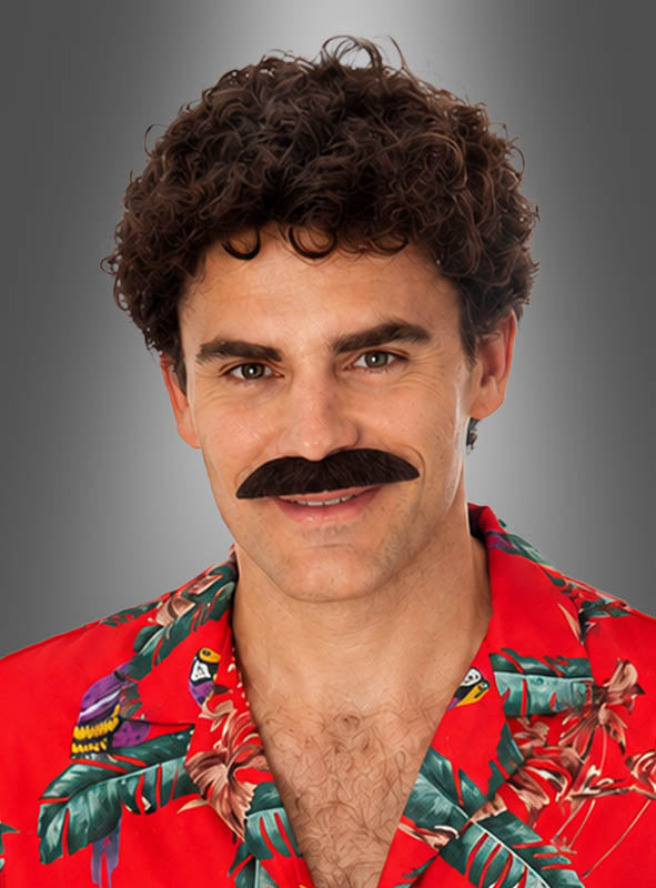Ein Mann mit lockigem braunem Haar und einem dichten, dunkelbraunen Bart im Magnum-Stil, der ein rotes Hawaiihemd mit tropischem Muster trägt, lächelt vor einem grauen Hintergrund mit Farbverlauf – ein Look, der direkt aus einem 80er-Jahre-Kostüm stammen könnte.