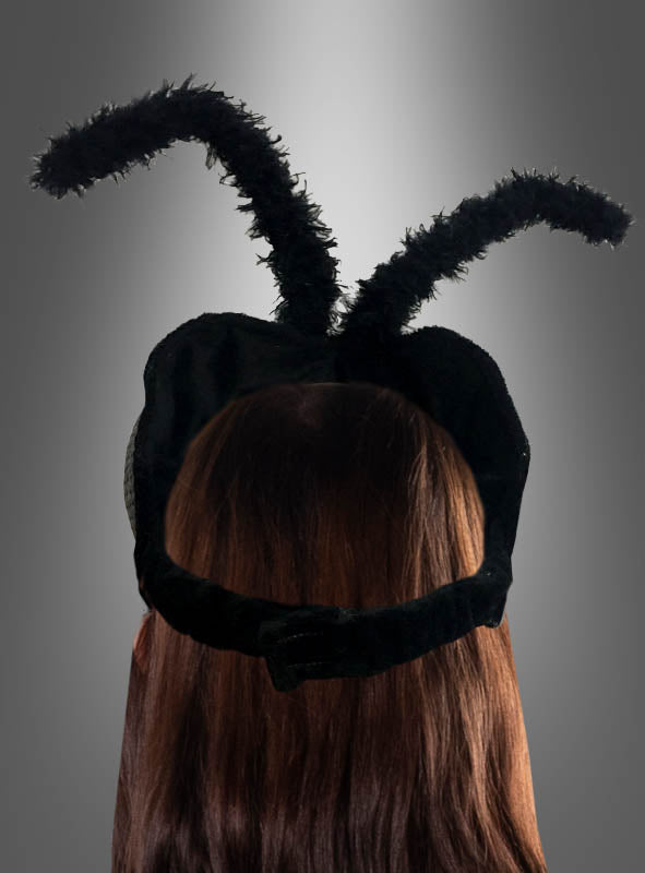 Eine Person mit langen braunen Haaren trägt ein schwarzes Stirnband mit der Aufschrift „Kopfteil Fliege Facettenaugen Insekt“ und zwei flauschigen Fühlern, die nach oben ragen und an die Facettenaugen eines Insekts erinnern, vor einem grauen Hintergrund mit Farbverlauf.