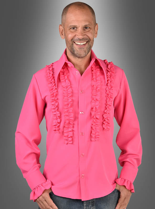 Ein lächelnder Mann trägt das „Waschbares Rüschenhemd 70er Jahre“ in leuchtendem Pink mit Rüschenverzierungen an Brust und Ärmelbündchen, während er vor einem grauen Hintergrund mit Farbverlauf steht – ideal für jede Schlagerparty oder als lustiges 70er-Jahre-Kostüm.