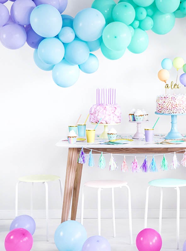 Hochwertige Luftballons in Pastellfarben schmücken einen Partytisch mit Kuchen, Bechern, Tellern und Hockern. Auf dem Boden liegen verstreut Luftballons, während vom Tisch eine Quastengirlande herabhängt.