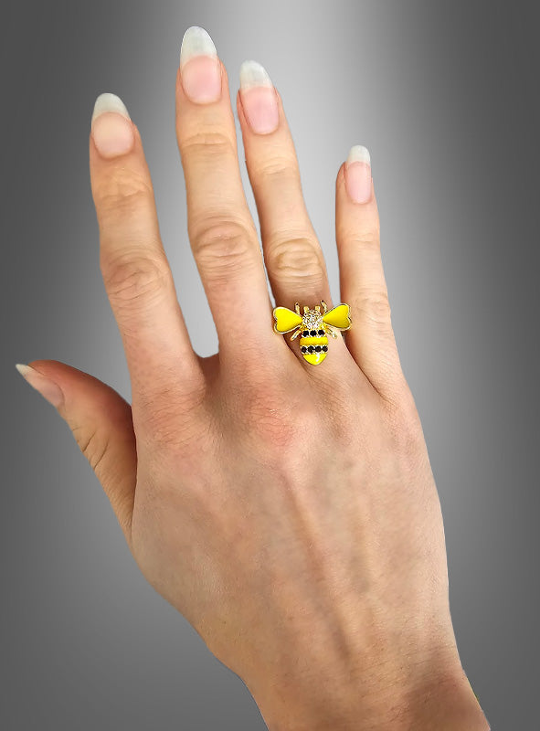 Eine Hand mit langen Naturnägeln trägt am Mittelfinger einen verstellbaren Bienenring mit Strasssteinen in Form einer Biene vor einem grauen Farbverlaufshintergrund – perfekt, um Ihrem Look eine verspielte Note mit Bienenkostüm-Schmuck zu verleihen.