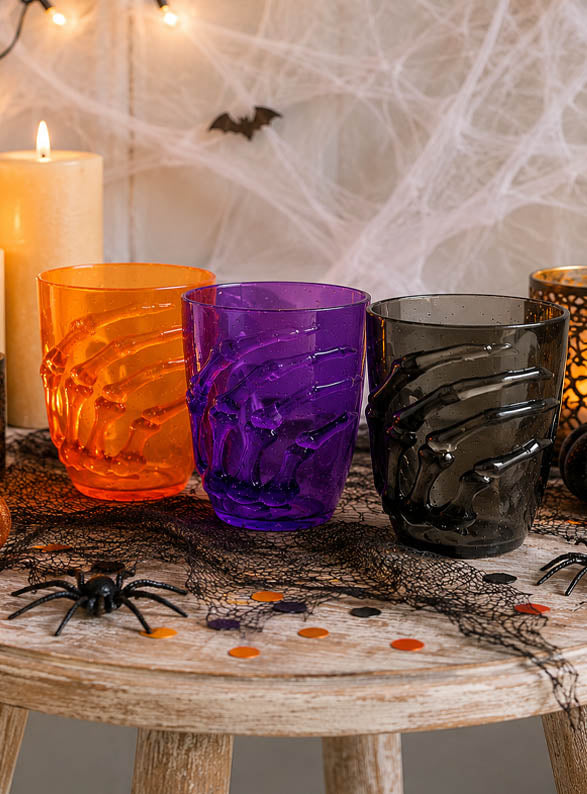 Drei „Halloween Glas“-Becher (10 cm) mit Skelett-Motiv – in Orange, Lila und Schwarz – stehen auf einem Holztisch, der mit künstlichen Spinnen, Kerzen, Konfetti und Spinnweben geschmückt ist und so die perfekte „Halloween Glas“-Tischdekoration bildet.