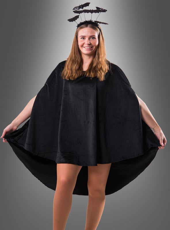 Eine Frau, die einen „Wandelbaren schwarzen Poncho aus Plüsch“ in den Größen XS–XXXL und ein Stirnband mit schwarzen Fledermausverzierungen trägt und vor einem grauen Hintergrund mit Farbverlauf steht – perfekt für dein nächstes Karnevalskostüm.