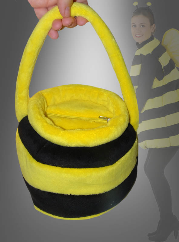 Eine Hand hält eine flauschige, runde „Biene Handtasche Honigtopf“ in Schwarz-Gelb mit gelben und schwarzen Streifen und einem Reißverschluss. Im Hintergrund ist teilweise eine Person in einem Bienenkostüm zu sehen, was dieses Accessoire zum perfekten Begleiter für jedes Bienenkostüm macht.