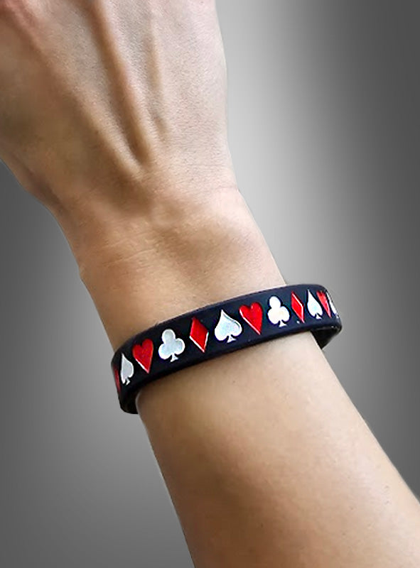 Eine Person trägt das „Casino Armband schwarz 1 Stück“ – ein schwarzes Silikonarmband mit roten Herzen und Rauten sowie weißen Pik- und Kreuz-Symbolen im klassischen Spielkartendesign – vor einem Hintergrund mit Farbverlauf.