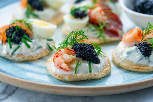 Auf einem blauen Teller sind kleine Blinis angerichtet, die mit Sahne, Kräutern, schwarzem Kaviar, Garnelen und in Scheiben geschnittenem gekochtem Ei belegt sind.