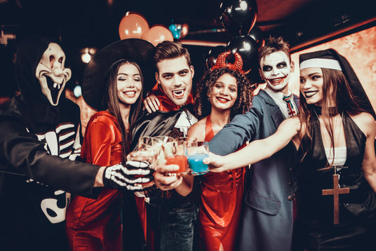 Sechs Personen in Halloween-Kostümen lächeln und stoßen auf einer Party gemeinsam an; im Hintergrund sind Dekorationen und Luftballons zu sehen.