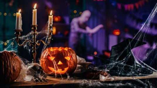 Eine geschnitzte Kürbislaterne, Kerzen und Spinnweben schmücken einen Tisch. Im Hintergrund steht eine verschwommene Gestalt, hinter der Halloween-Dekorationen zu sehen sind.