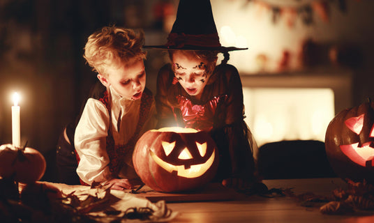 Zwei Kinder in Halloween-Kostümen betrachten eine brennende Kürbislaterne auf einem Tisch, umgeben von Kerzen und Herbstdekorationen in einem schwach beleuchteten Raum.