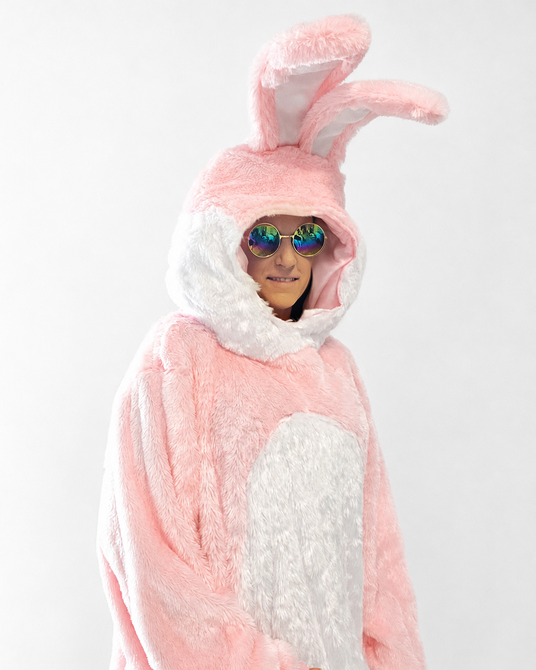 Eine Person in einem flauschigen rosa-weißen Hasenkostüm mit großen Ohren und einer runden, verspiegelten Sonnenbrille, die vor einem schlichten weißen Hintergrund steht.