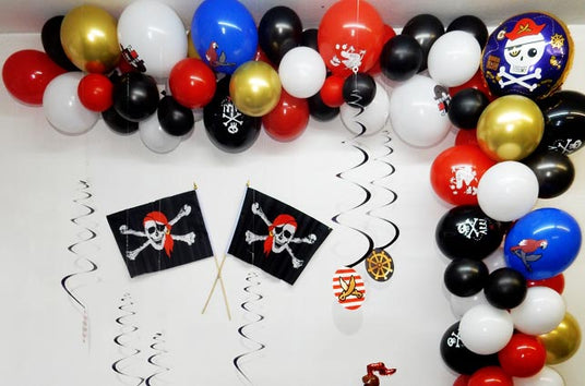 Partydekoration im Piratenstil mit schwarzen, roten, weißen und goldenen Luftballons, Piratenflaggen und spiralförmigen Luftschlangen an einer weißen Wand.
