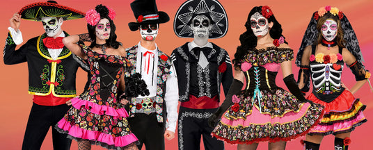 Sechs Menschen in farbenfrohen „Día de los Muertos“-Kostümen und mit Gesichtsbemalung stehen nebeneinander vor einem rosa-orangefarbenen Hintergrund.