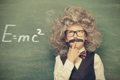 Ein Kind, das als Wissenschaftler verkleidet ist und einen falschen Schnurrbart sowie eine Perücke trägt, steht vor einer Tafel, auf der „E=mc²“ steht, und blickt nachdenklich nach oben.