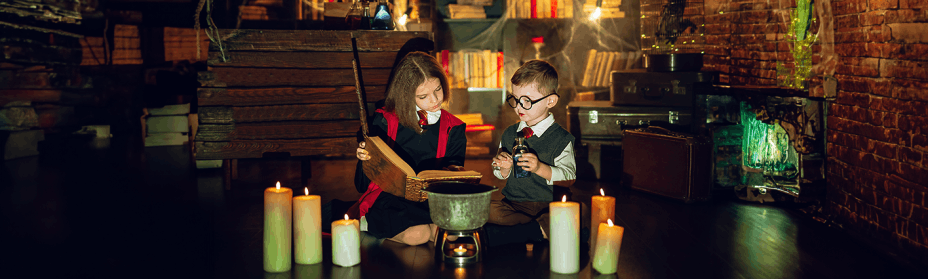 Zwei Kinder in Kostümen sitzen, umgeben von Kerzen, in einem schwach beleuchteten Raum voller alter Bücher und geheimnisvoller Gegenstände, lesen in einem großen Buch und halten Fläschchen in den Händen.