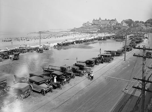 Schwarz-Weiß-Foto von Oldtimern, die entlang einer Strandstraße geparkt sind, mit Menschen im Sand und einem großen Hotel auf einem Hügel in der Ferne.