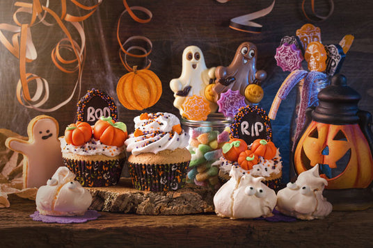 Desserts zum Thema Halloween, darunter Cupcakes mit „BOO“- und „RIP“-Verzierungen, Leckereien in Geisterform, Kürbisdekorationen, Plätzchen und ein Glas mit einer Jack-o-Lantern auf einem Holztisch.