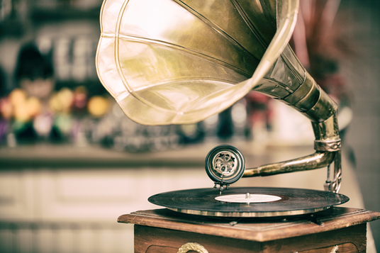 Ein Vintage-Grammophon mit einem großen Messingtrichter spielt eine Schallplatte auf einem Holzsockel ab; im Hintergrund ist ein verschwommener Hintergrund zu sehen.
